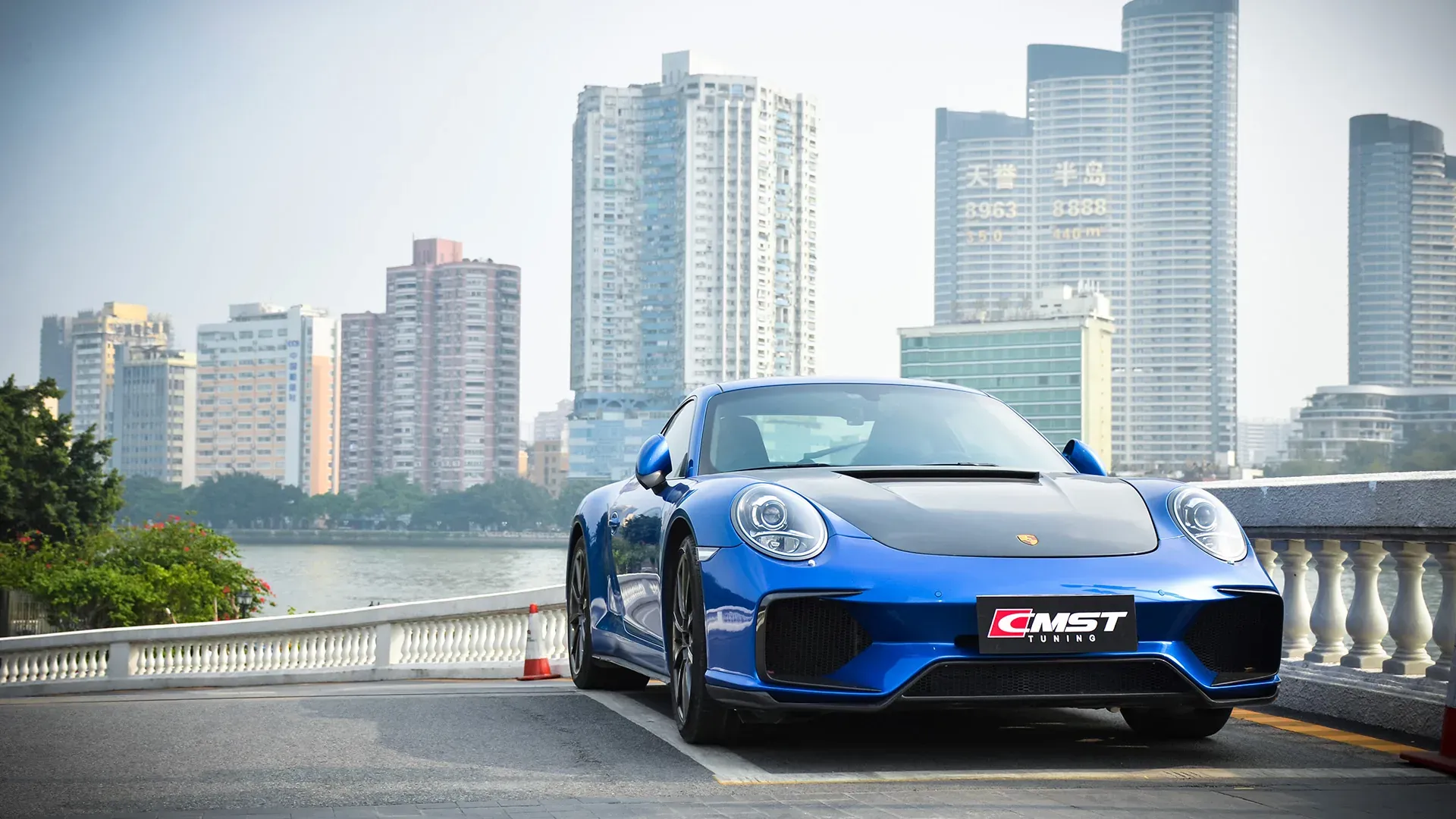 Blue Porsche 911 photographed from the front three-quarter, with a CMST front bumper and front lip fitted, on a bridge against a city skyline
