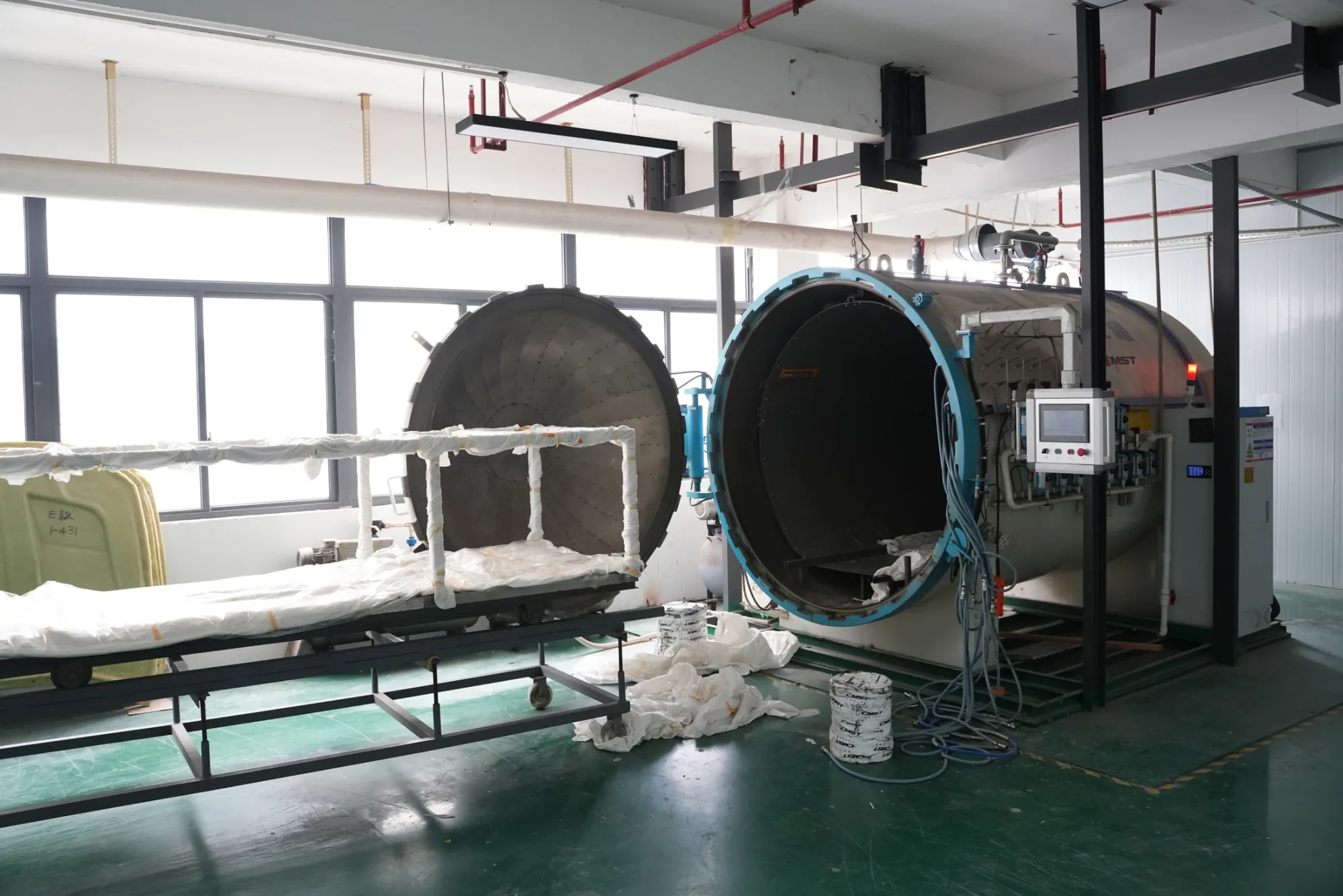 Vacuum-bagged carbon parts on a trolley beside two curing vessels in a composites workshop